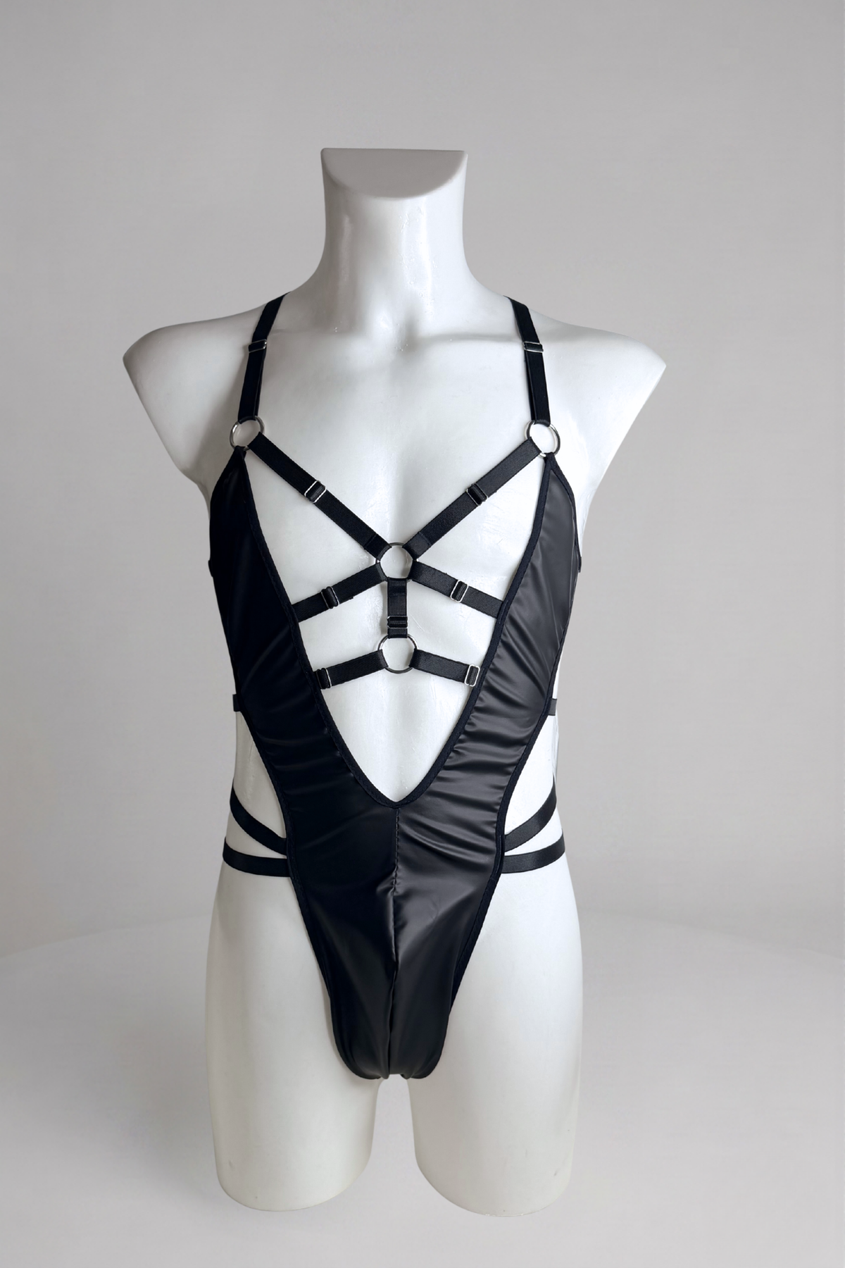 Black vegan leather harness bodysuit for men on mannequin, front view: deep V with O-ring chest straps