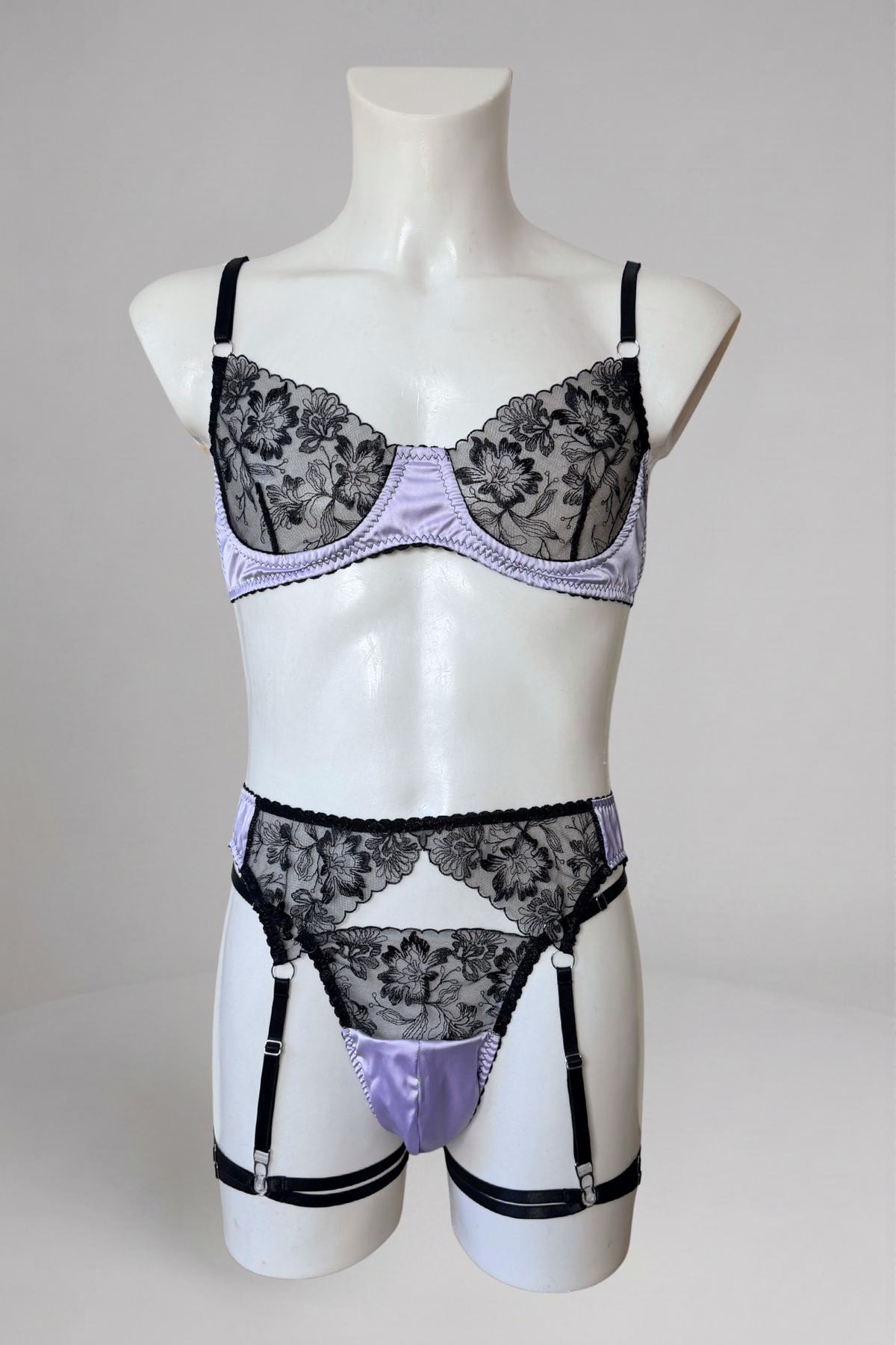 Ensemble lingerie homme en satin lavande sur mannequin, vue de face : soutien-gorge en dentelle noire, string en satin et porte-jarretelles