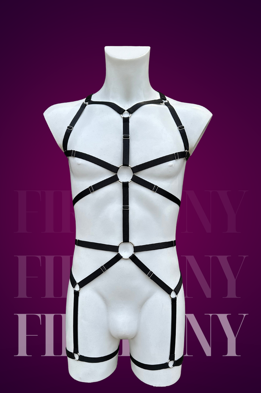 Full-body black strappy harness for men with silver rings, adjustable elastic design, displayed on a mannequin against a purple background.

